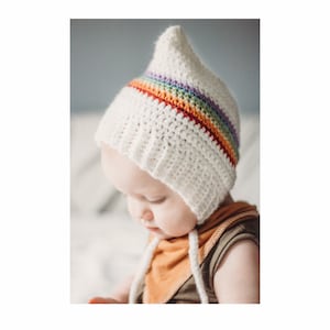 Puede incluir: Un gorro de bebé de crochet blanco con una franja de arcoíris. El gorro tiene una parte superior puntiaguda y se ata debajo de la barbilla.
