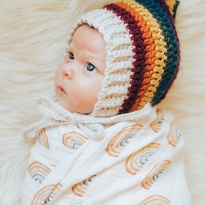Puede incluir: Un bebé con un gorro de crochet de arcoíris y envuelto en una manta de muselina blanca con un patrón de arcoíris.