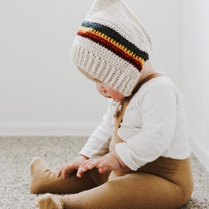Puede incluir: Un bebé con un gorro de crochet blanco con una franja de arcoíris y polainas marrones. El bebé está sentado en una alfombra.