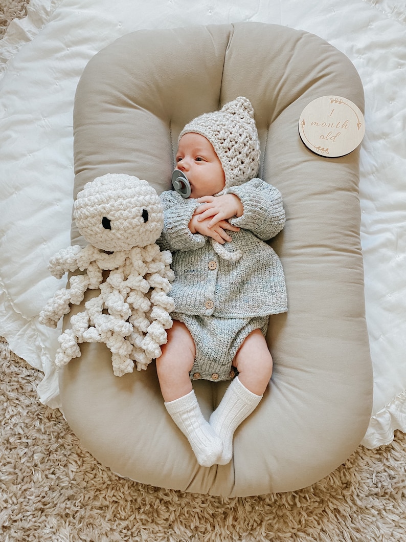 Puede incluir: Un beb&eacute; con un gorro de crochet blanco y un c&aacute;rdigan de punto gris est&aacute; acostado en un nido beige. El beb&eacute; sostiene un juguete de pulpo de crochet blanco y tiene un chupete en la boca. Un letrero de madera con el texto "1 mes" es visible en el fondo.