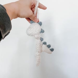 Könnte beinhalten: Weißes gehäkeltes Dinosaurier-Ornament mit blauen Akzenten. Der Dinosaurier hat einen langen Schwanz und einen kleinen Kopf mit einer spitzen Schnauze. Das Ornament hängt an einer weißen Schnur.