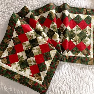 May include: A rectangular quilted table runner featuring a patchwork design of red, dark green, and cream-colored fabric squares. The pattern includes printed motifs of holly leaves, red berries, and pinecones, finished with a wide green border.