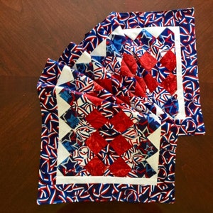 May include: Red, white, and blue quilted table runner with a star and ribbon pattern. The runner has a white border and is folded in half.