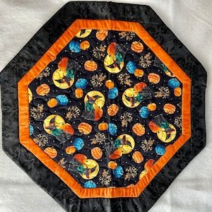 May include: An octagonal quilted table topper featuring a black fabric center with a pattern of black cats wearing witch hats, orange and blue pumpkins, and yellow moons. The center is framed by an orange border and a wide black damask border.