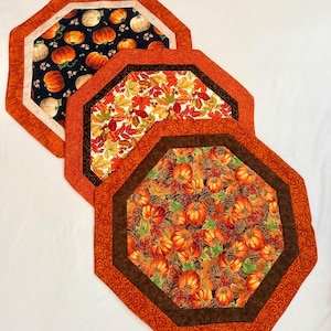 May include: Three octagonal fabric placemats featuring autumn-themed patterns. One has pumpkins on a black background, one displays colorful fall leaves, and the third shows pumpkins with pinecones. Each has a wide orange or brown border.