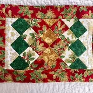 May include: A rectangular quilted placemat featuring a patchwork design of diamond-shaped fabric pieces in red, dark green, and gold. The fabric patterns include holly leaves, berries, and pine needles. A wide red border frames the central pattern.
