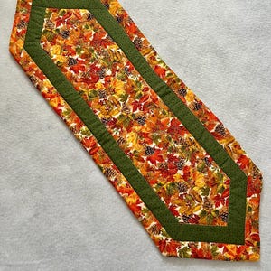 May include: A decorative table runner featuring a fall-themed pattern. The runner has a central design of autumn leaves and pinecones in shades of red, orange, and yellow. It is bordered by a green fabric with a dotted pattern.