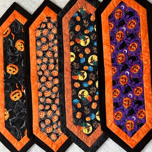 May include: Four Halloween-themed table runners. Each runner features a black border with an orange inner border. The center fabric displays various Halloween patterns, including pumpkins, cats, and witch designs. The runners are likely made of fabric.