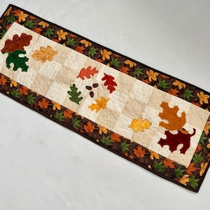 May include: A rectangular quilted table runner featuring a central panel of alternating cream and beige squares. The border is dark brown with a pattern of small green, orange, and yellow leaves. Appliqué oak leaves in red, green, and gold accent the runner.