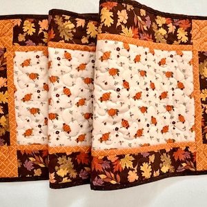 May include: A quilted table runner featuring a central cream panel with an orange pumpkin print, bordered by a dark brown fabric with a colorful autumn leaf pattern. The runner includes orange dotted fabric accents and decorative stitching.