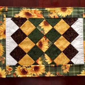 May include: Rectangular quilted placemat featuring a sunflower and plaid design. The border showcases large yellow sunflowers on a green plaid background. The center has a diamond pattern in yellow, brown, and green.