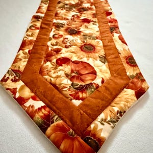 May include: A quilted table runner featuring a central panel with a print of sunflowers, pumpkins, and autumn leaves on a cream background, framed by a solid burnt orange border with mitered corners and a pointed end design.