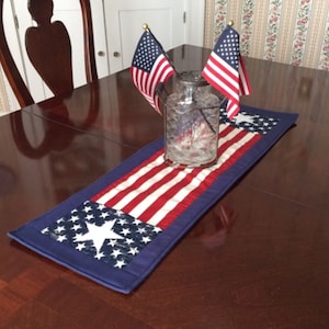 May include: A blue and red table runner with a star pattern, featuring a glass vase with two small American flags. The runner is on a dark wood table.