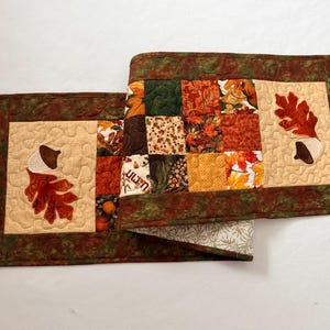 May include: A rectangular quilted table runner featuring a patchwork center of autumn-themed fabric squares in shades of orange, yellow, brown, and green. The ends are cream-colored panels with appliqued oak leaves and acorns, framed by a dark border.
