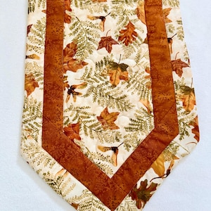May include: A quilted table runner featuring a cream-colored center panel with a pattern of green ferns, brown oak leaves, and maple seeds. The center is framed by solid, textured rust-brown fabric borders, with matching leaf-patterned fabric edges.