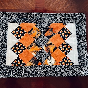 May include: A rectangular quilted placemat featuring a central patchwork design of orange fabric squares and black fabric squares with candy corn patterns. The center is framed by white fabric strips and a border of black fabric with white spiderwebs.
