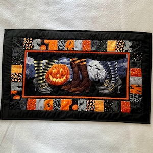 May include: A rectangular Halloween-themed quilted table runner. The design features a carved pumpkin, witch boots, and a spooky background. The border is black with orange and black patterned squares. The overall color scheme is orange, black, and gray.