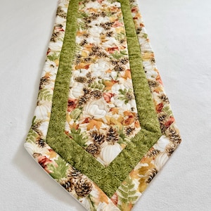 May include: A quilted table runner featuring a central panel with a pattern of white pumpkins, brown pinecones, and autumn leaves in shades of orange, yellow, and green, bordered by a textured olive green fabric with a subtle leaf print design.
