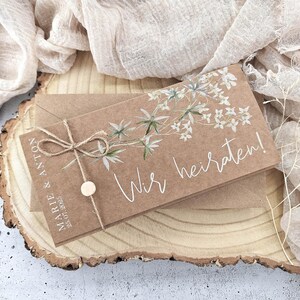 May include: A brown card with white floral illustrations and the text "Wir heiraten!"  The card is tied with twine and has a small wooden circle attached. The card is sitting on a wooden surface.