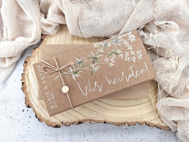 May include: A brown card with white floral illustrations and the text "Wir heiraten!"  The card is tied with twine and has a small wooden circle attached. The card is sitting on a wooden surface.