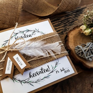 K&ouml;nnte beinhalten: Eine rustikale Hochzeitseinladung mit einem Burlappsband und einer wei&szlig;en Feder. Die Einladung ist mit einem Kranz aus gr&uuml;nen Bl&auml;ttern und dem Text "Wir heiraten!" und "25. Juli 2018" verziert.