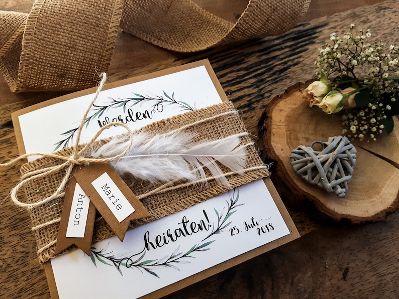 K&ouml;nnte beinhalten: Eine rustikale Hochzeitseinladung mit einem Burlappsband und einer wei&szlig;en Feder. Die Einladung ist mit einem Kranz aus gr&uuml;nen Bl&auml;ttern und dem Text "Wir heiraten!" und "25. Juli 2018" verziert.