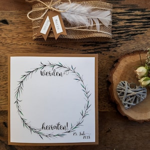 K&ouml;nnte beinhalten: Eine braune Karte mit wei&szlig;em Hintergrund und einem gr&uuml;nen Kranz-Design. Der Text "werden" und "heiraten!" ist in schwarzer Kalligraphie geschrieben. Das Datum "25. Juli 2018" ist in Schwarz unter dem Kranz geschrieben.