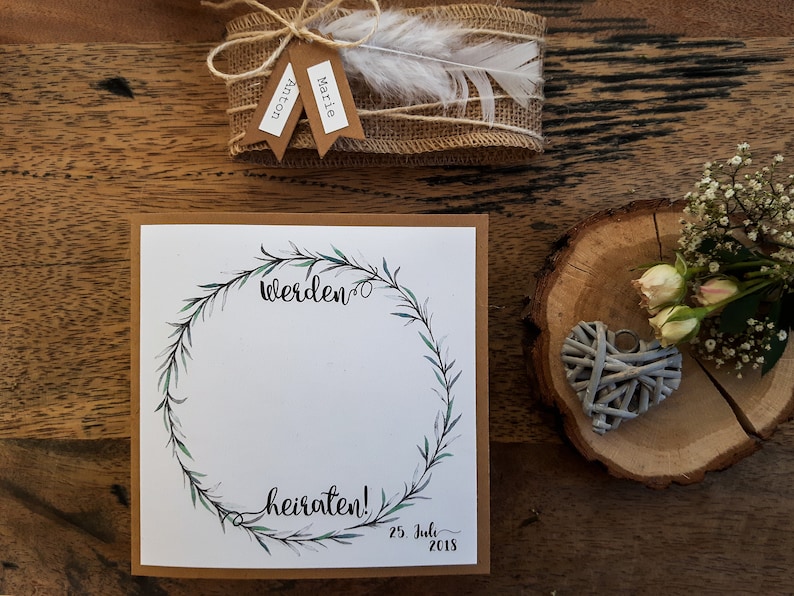 K&ouml;nnte beinhalten: Eine braune Karte mit wei&szlig;em Hintergrund und einem gr&uuml;nen Kranz-Design. Der Text "werden" und "heiraten!" ist in schwarzer Kalligraphie geschrieben. Das Datum "25. Juli 2018" ist in Schwarz unter dem Kranz geschrieben.