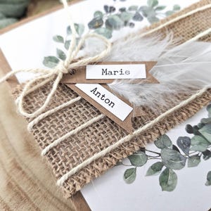 May include: Two rustic place cards with the names "Marie" and "Anton" written on them. The cards are tied together with twine and decorated with a white feather and greenery.