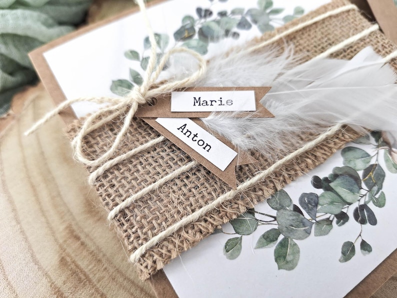 May include: Two rustic place cards with the names "Marie" and "Anton" written on them. The cards are tied together with twine and decorated with a white feather and greenery.
