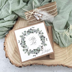 May include: A white card with a green watercolor wreath and the text "All the Love" in black calligraphy. The card is on a brown wooden slice with a brown envelope and a burlap gift box with a white feather.