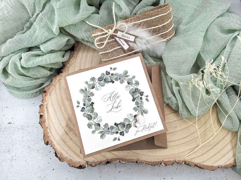 May include: A white card with a green watercolor wreath and the text "All the Love" in black calligraphy. The card is on a brown wooden slice with a brown envelope and a burlap gift box with a white feather.
