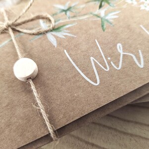 May include: A brown paper card with a white floral design and the text "Wir he" in white script. The card is tied with twine and a white wooden bead.