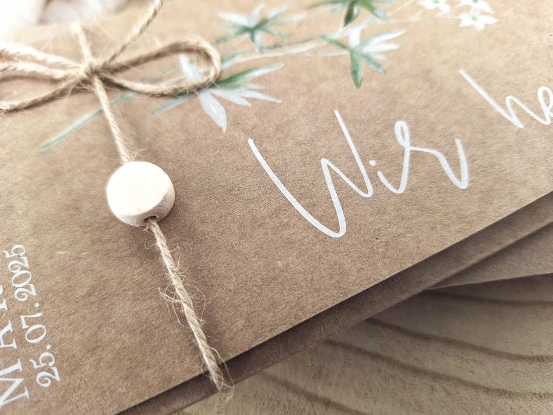 May include: A brown paper card with a white floral design and the text "Wir he" in white script. The card is tied with twine and a white wooden bead.