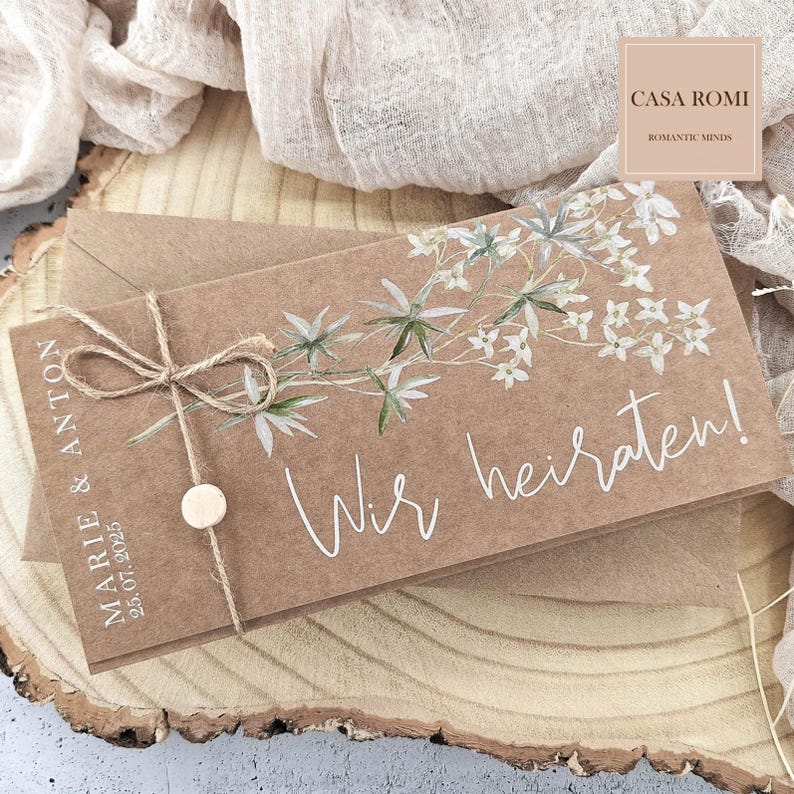 May include: A brown card with white floral designs and the text "Wir heiraten!"  The card is tied with twine and has a small wooden circle attached. The text "MARIE & ANTON 25.07.2025" is printed on the left side of the card. The text "CASA ROMI ROMANTIC MINDS" is printed in the top right corner of the card.