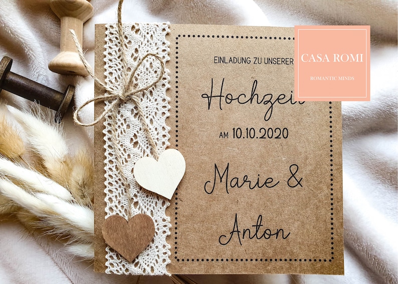 May include: A brown paper wedding invitation with a lace trim and wooden hearts. The invitation reads "Einladung zu unserer Hochzeit am 10.10.2020 Marie & Anton" and has a logo that says "Casa Romi Romantic Minds".