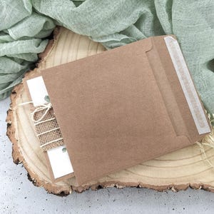 May include: A brown kraft paper envelope with a white card inside. The envelope has a white paper seal on the flap. The envelope is sitting on a wooden slice with a piece of burlap fabric tied around it.