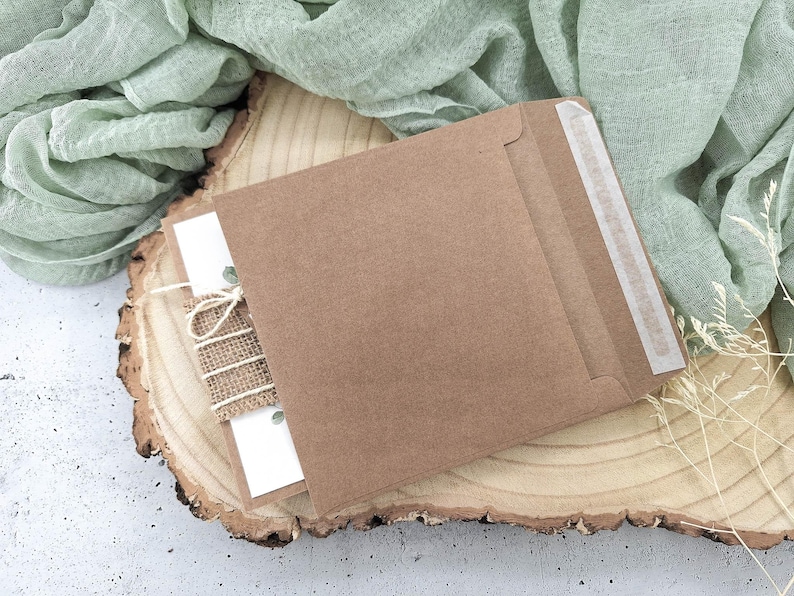 May include: A brown kraft paper envelope with a white card inside. The envelope has a white paper seal on the flap. The envelope is sitting on a wooden slice with a piece of burlap fabric tied around it.