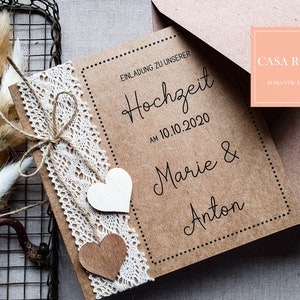 May include: A brown kraft paper wedding invitation with white lace trim and two wooden hearts. The invitation reads "Einladung zu unserer Hochzeit am 10.10.2020 Marie & Anton" and is decorated with a brown twine bow. The invitation is on a white background.