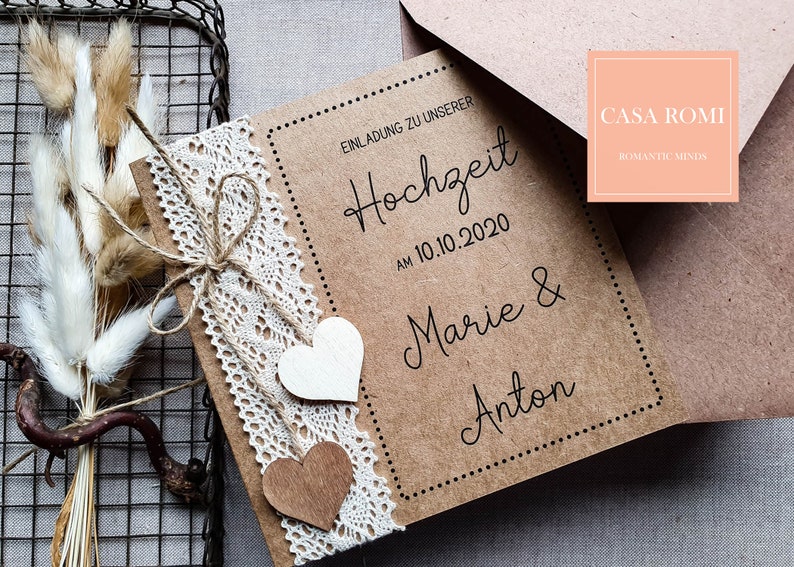 May include: A brown kraft paper wedding invitation with white lace trim and two wooden hearts. The invitation reads "Einladung zu unserer Hochzeit am 10.10.2020 Marie & Anton" and is decorated with a brown twine bow. The invitation is on a white background.