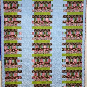 May include: A quilt with three vertical panels of stacked fabric strips. The strips are in various patterns and colors, including pink, green, brown, and blue. The background is a light blue.