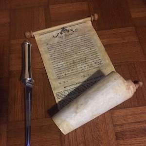 Puede incluir: Una daga plateada y azul con un pergamino. La daga tiene un mango detallado y una hoja larga y puntiaguda. El pergamino est&aacute; hecho de papel envejecido y tiene texto. El pergamino est&aacute; parcialmente enrollado, y la daga est&aacute; al lado sobre una superficie de madera.