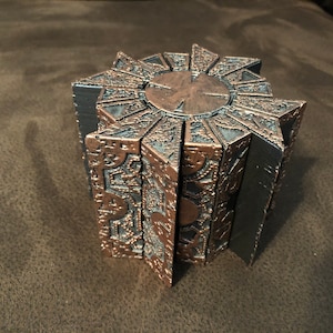 May include: A bronze-colored puzzle box with intricate geometric patterns and a central circular design. The box is composed of multiple panels that appear to be partially open, revealing a dark interior. The overall aesthetic suggests a complex, possibly supernatural, object.