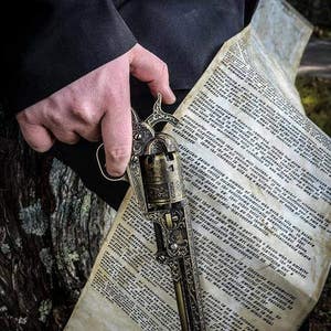 Puede incluir: Un rev&oacute;lver ornamentado de estilo antiguo con intrincados grabados, sostenido sobre un pergamino con texto y un s&iacute;mbolo de pentagrama. El arma tiene un acabado de bronce y el pergamino tiene tonos de papel envejecido.