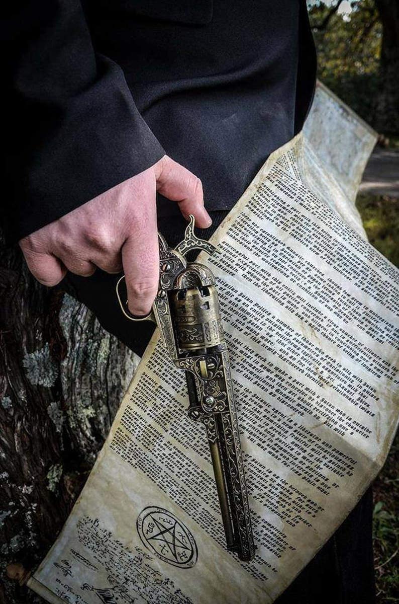 Puede incluir: Un rev&oacute;lver ornamentado de estilo antiguo con intrincados grabados, sostenido sobre un pergamino con texto y un s&iacute;mbolo de pentagrama. El arma tiene un acabado de bronce y el pergamino tiene tonos de papel envejecido.