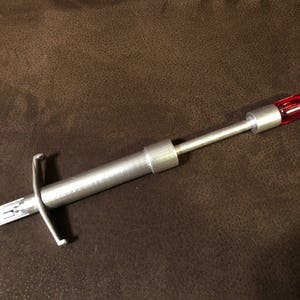 Puede incluir: Una herramienta plateada con una punta roja rectangular. La herramienta tiene un cuerpo largo y cilíndrico con una estructura similar a un mango en un extremo y una pieza en forma de cruz cerca del otro. El objeto está sobre una superficie marrón.