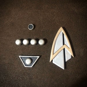 May include: A collection of Star Trek-themed metal pins and emblems. The set includes a silver and gold arrowhead-shaped emblem, a black and silver triangle, four small silver spheres, and a small silver cylinder.
