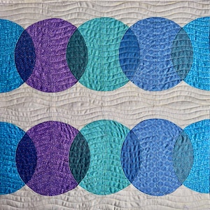 May include: A quilted textile featuring a repeating pattern of overlapping circles in shades of purple, teal, and blue. The circles are set against a neutral, wavy background, creating a modern, geometric design. The quilt is machine-stitched.
