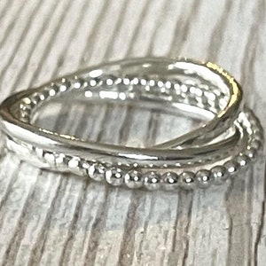 May include: A silver ring featuring three interlocking bands. One band has a textured finish, another is adorned with small beads, and the third is smooth. The ring is presented on a light-coloured wooden surface.