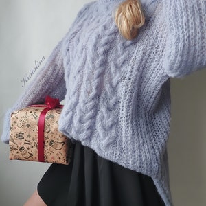 Peut inclure: Un pull en maille bleu clair avec un motif en tricot torsad&eacute;. Le pull est ample et a une forme longue et fluide. Le pull est port&eacute; par une personne avec une jupe noire et des collants marrons.